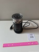 Nespresso Aeroccino milk frother with power cord on white surface, showing entire unit with lid attached on side and pink ruler for scale.