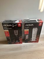 Two boxed NOMA heaters, black digital oil-filled heater and white oil-filled heater, standing side by side on floor.