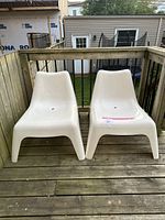 Two white plastic outdoor chairs placed side by side on a wooden deck with railing background and yard beyond.