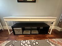 White Ikea Micke desk with two drawers shown from front, placed on a patterned rug and hardwood floor with a black storage unit underneath containing fabric bins.