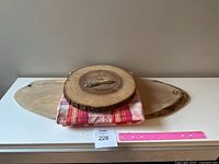 Front view of two large wooden serving boards stacked with a folded red and white checkered tablecloth and a round wooden board on top, showing natural wood grain and bark edges, with a decorative rabbit cheese knife on top.