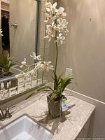 Artificial white orchid plant with multiple blooms and green leaves in gray vase on bathroom counter.