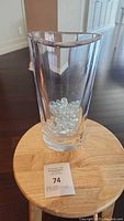 Front view of clear asymmetric pillar glass vase with a number of clear glass marbles inside, sitting on a wooden stool.