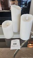 Three battery-operated flameless candles in white of varying sizes with LED flame tips on a glass table.