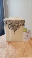 Front view of square glass tissue box case with intricate black floral and scroll design on cream background.