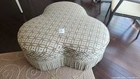 Top view of custom foot stool showing three-leaf clover shape, patterned fabric in white and gold, with fringe trim around base