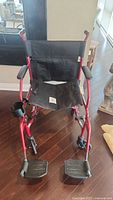 Front view of the foldable wheelchair showing black fabric seat and footrests, red frame, and armrests.