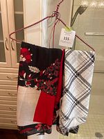 Two Gerry Weber scarves displayed on a pink and red wire hanger with tags attached. One scarf has a red, black, beige floral and geometric pattern, and the other has a black and white plaid/checkered pattern.