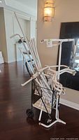 White metal garment rack parts loosely assembled in black mesh wire basket frame on wheels, leaning against wall indoors.