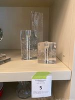 Three clear glass candle holders of varying heights and triangular prism shapes displayed on a shelf, each with visible embedded bubbles and a candle slot on top.