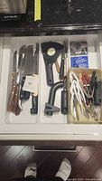 Top-down view of drawer showing knives on left, kitchen utensils including peeler, can opener, tongs on right, and box of storage bags