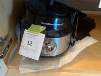 Hamilton Beach food processor base unit with control dial in stainless steel and black plastic finish, power cord visible, placed on a shelf with paper towel underneath.