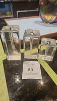 Three clear glass modernist single stem bud vases in block form, shown together on a countertop with auction lot tag 64 in front.