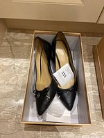 Pair of black leather stilettos with pointed toes and small decorative bows, placed inside a beige-tan shoe box.