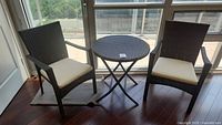 Wide angle view showing two dark brown woven armchairs with cream cushions and a matching round table with black metal folding legs near a window.