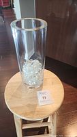 Clear cylindrical glass vase displayed on a wooden stool with clear decorative marbles inside, shown from the front.