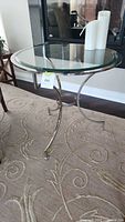 Side view of round glass top table with stainless steel curved legs and base support, placed on patterned carpet.