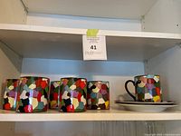 Shelf display showing six colorful mugs and two matching plates from the Dieci by Taitu collection.