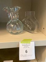 Two clear glass vases on a shelf; left one is a ruffled flint glass vase with vertical ribbing, right one is a flat Kosta Boda glass vase with floral design.