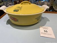 Yellow cast iron enamel saucepan with lid and colorful fruit motif on side, placed on a pale surface with auction lot card visible.