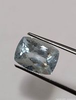 Close-up image of a single cushion cut light blue aquamarine gemstone held by tweezers showing facets and internal inclusions.