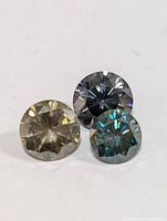 Three round faceted moissanite gemstones in light yellow, dark gray, and bluish-green colors displayed on a white background.