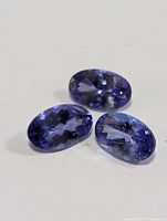 Close-up photo showing three oval-shaped faceted tanzanite gemstones with violet-blue color on a white background.
