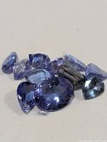 Close-up photo of loose faceted tanzanite gemstones in varying cuts and sizes, displaying their blue-violet hues and some chips or breaks.