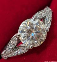 Close-up front view of silver ring with central round moissanite stone on a red background, showing detailed milgrain work.