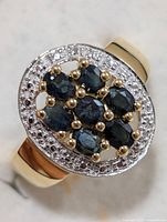 Close-up angle of oval cluster ring featuring seven oval sapphires and surrounding halo of small white stones, showing gold-tone prong settings on a silver band.