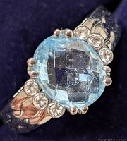 Close-up photo showing the oval faceted blue topaz at the center of the sterling silver ring, with three small clear stones on each side and engraved band details.