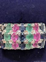 Close-up image showing the front view of a wide sterling silver ring with an alternating pattern of oval emerald, sapphire, and ruby gemstones in multiple rows.