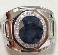 Close-up photo of silver ring with a large oval blue sapphire surrounded by small clear stones, showing top view of the ring.