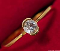 Close-up photo of the silver ring featuring a clear round faceted stone set in a yellowish prong setting, shown on a red background.