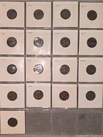Top-down photo of 17 Canadian pennies from the 1950s individually secured in white cardboard holders arranged in a 4x4 grid with an extra holder empty at the bottom.