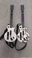 Two Guitar Hero guitar controllers shown front side with one decorated with several cartoon-style stickers and colorful fret buttons on neck.