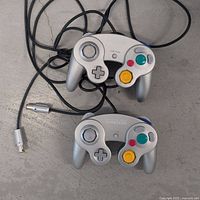Front view of two silver Nintendo GameCube controllers showing button layouts and cables.