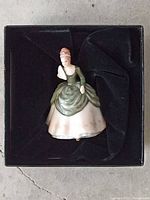 Miniature lady statue in factory packaging, showing front view, dressed in green and pink period gown with floral accents.