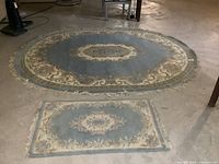 Wide view of the large oval wool area rug placed on a concrete floor with smaller matching rectangular rug in front of it showing design and size complement.