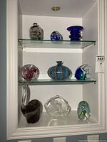 View of all art glass items arranged on two glass shelves including vases, paperweights, and sculptural pieces showing their shapes and colors