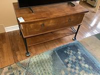 View of the cabinet front showing wood finish, drawer with two metal ring handles, and metal base with feet on hardwood floor.