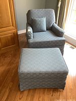 Blue patterned upholstered arm chair with square throw pillow, matching ottoman in front, hardwood floor and cabinet in background