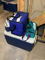 Wide view showing the three coolers stacked in basement. Large blue hard cooler at bottom, medium blue soft cooler on top, green soft cooler on top right.