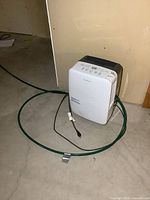White Garrison dehumidifier with attached green garden hose on basement floor.