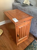 Side view of the wooden end table showing the paneled cabinet door with brass handle and slide-out shelf knob on the top edge.