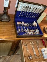 Open wooden chest containing Community brand silverplate knives, forks, spoons arranged in blue velvet-lined slots with additional serving pieces and napkin rings below in drawer.