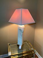 Full view of the lamp lit, showing the white ram head base and light fabric lampshade on a glass-top table.