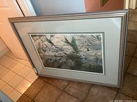 Full view of the framed print leaning against a wall on a tile floor, showing the overall size and framing style.