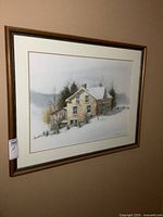 Framed print of a stone house in winter with snow, wooden fence and bare trees visible. The print is behind glass in a wooden frame.