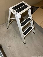 White metal folding step stool with three steps showing side angle view on concrete floor near cardboard box.